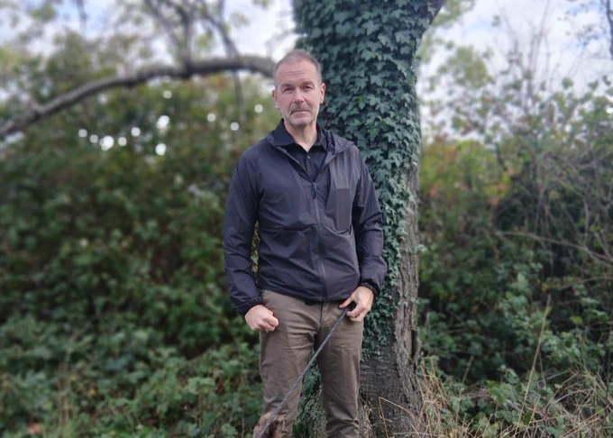 Man wearing a black jacket and khaki pants standing outdoors near a tree covered in ivy with dense greenery in the background.