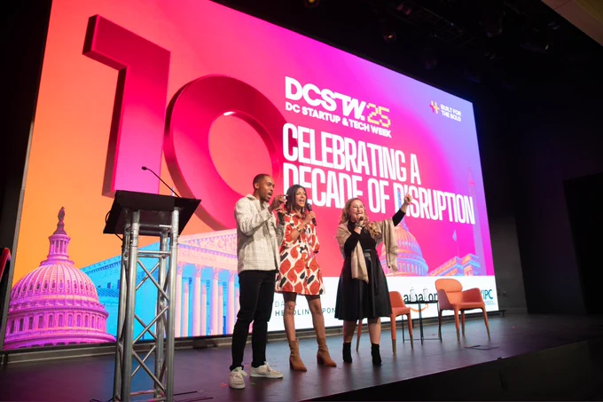 Three people stand on stage speaking into microphones at DC Startup Week, with a large screen behind them reading "Celebrating a Decade of Disruption.