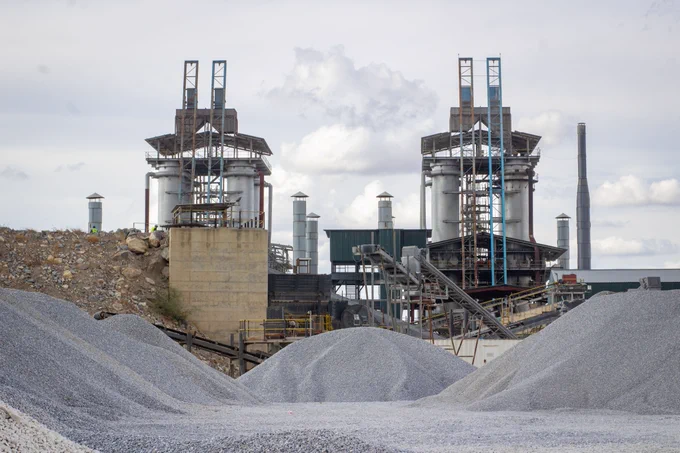 Industrial site with stockpiles in the foreground. Two large processing towers are central, flanked by additional industrial structures. The sky is overcast.