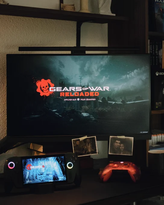 Setup gaming avec un écran affichant Gears of War: Reloaded, une ROG Xbox Ally X montrant Gears 5, une manette Xbox rouge et des photos polaroid de personnages du jeu posées sur le bureau.