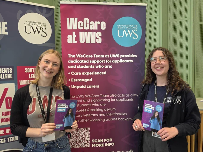 Two individuals are standing in front of a University of the West of Scotland (UWS) banner that reads "WeCare at UWS." They are holding promotional materials. The banner highlights support services for students, including care-experienced and estranged students, and unpaid carers.