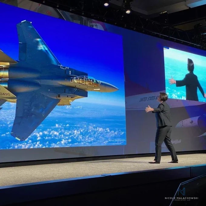 Nicole Malachowski stands on a large stage with arms open toward a giant screen displaying a fighter jet flying above the clouds. The stage presentation appears aviation or military themed, with dramatic blue lighting and a second screen showing the speaker from behind. The logo for Nicole Malachowski & Associates appears in the bottom-right corner.