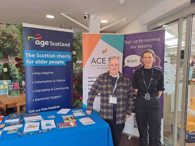 Man and woman at a table with charity banners for Age Scotland and ACE, promoting scam awareness.