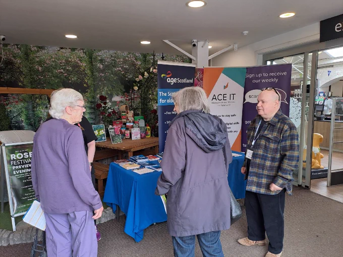 Three people at a stall promoting Age Scotland and ACE IT, with a floral backdrop and display tables.
