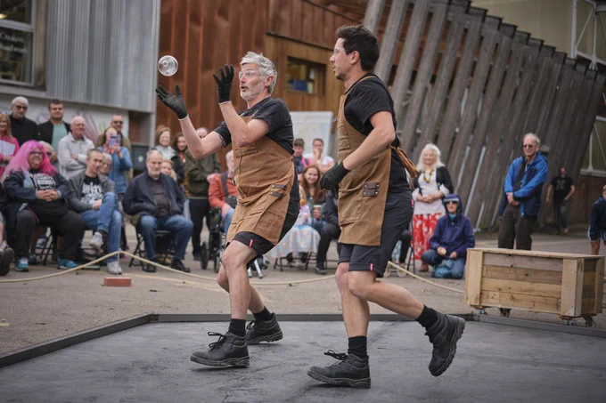 A street performance scene with two individuals in brown aprons juggling. An audience seated and standing in the background watches the act. The setting appears to be an outdoor, industrial-style area with a rusty metal building.