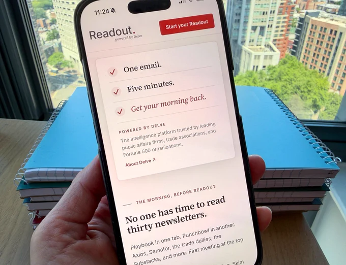 A hand holds a smartphone displaying a newsletter app called Readout with a cityscape and stacked notebooks visible in the background.