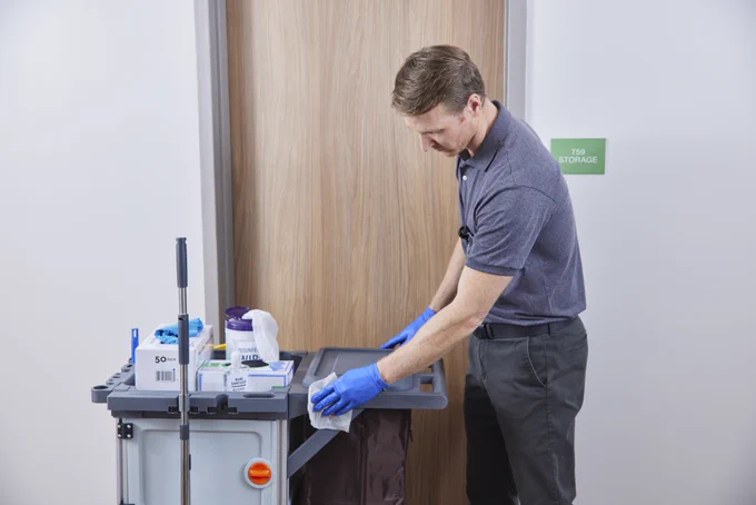 EVS worker wearing blue gloves and gray scrubs is wiping cart in a hallway.