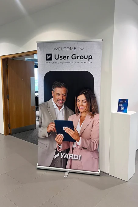 A banner in a hallway features two smiling individuals looking at a tablet. The banner reads "Welcome to User Group: Knowledge, Networking & Interaction" and displays the Yardi logo. In the background, there's a partially open wooden door.
