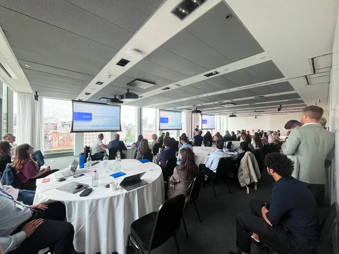 A conference room filled with attendees seated at round tables, facing multiple presentation screens displaying a blue background. Large windows show a cityscape view.