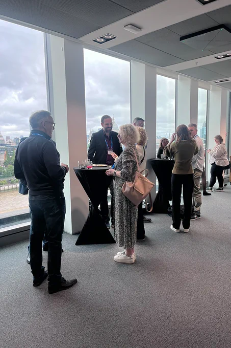 A group of people are gathered around tall tables in a modern conference room with large windows. Cityscape views are visible outside. The atmosphere appears to be one of networking or casual conversation.