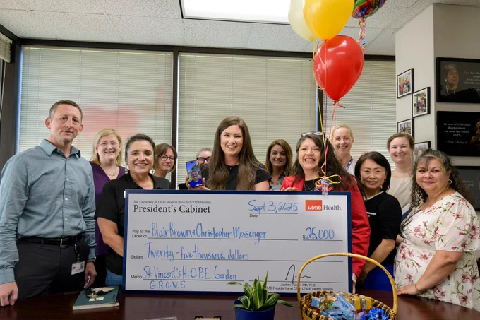A group of people smiling in an office setting, holding a large ceremonial check for $25,000 from UMC Health. The check is written to Blair Brown and Christopher Messenger for "St. Vincent's H.O.P.E. Garden." Some individuals are holding balloons.
