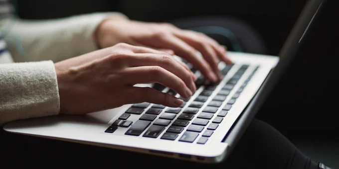 A person wearing a gray sweater types on a silver laptop with black keys.