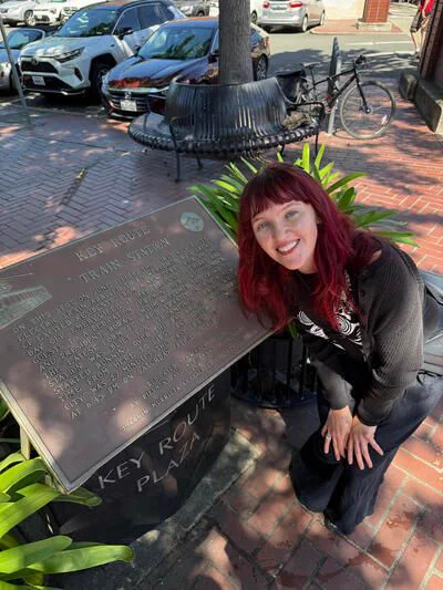 Kyra Eyerman posing beside the Key Route Train Station plaque in Oakland's Key Route Plaza. The plaque commemorates the arrival of the first Key Route electric train in 1904 and the station's role in East Bay transportation until the line's final run in 1958.