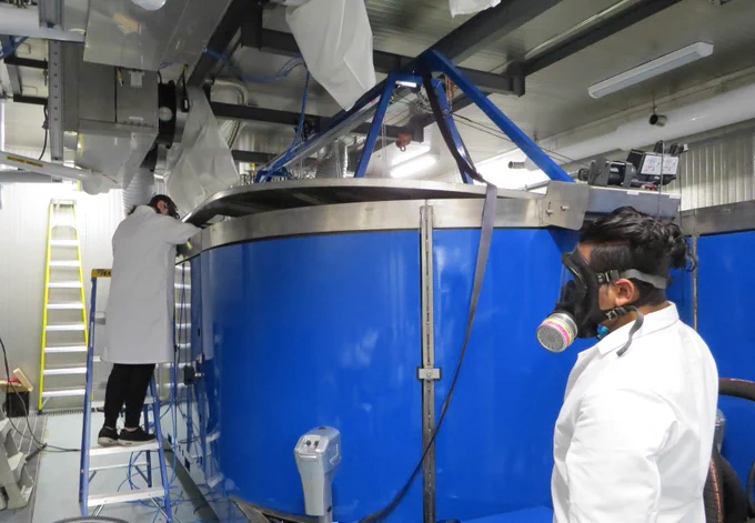 Scientists wearing white lab coats and respirators stand beside the large blue simulator tank. 