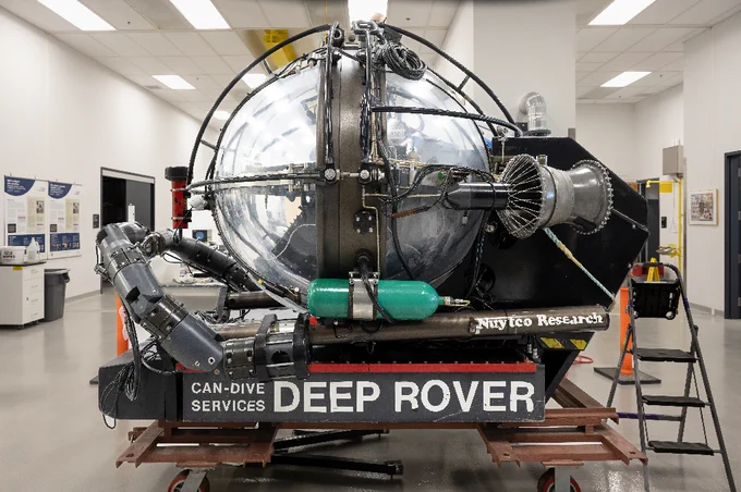 Submersible sphérique en eaux profondes étiqueté « Deep Rover » à l’intérieur d’une installation de recherche avec des bras mécaniques et de l'équipement attaché.