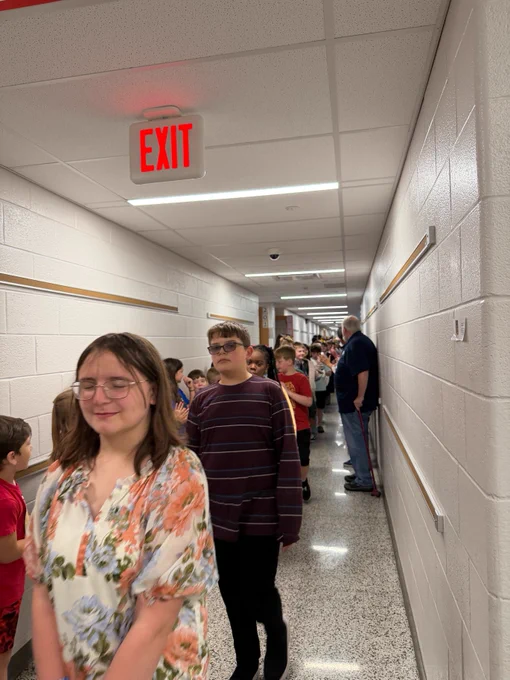 A group of students walking in a single-file line down a school hallway, guided by an adult. 