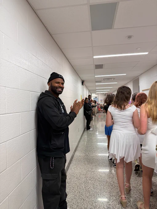 A person stands against a hallway wall, smiling and clapping, while a group of people in formal attire walk along the corridor.