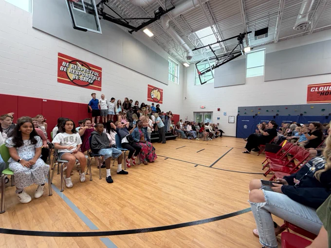 A group of people, mostly younger individuals, are gathered in a gymnasium. Some are sitting on chairs while others stand on a platform against the wall. 