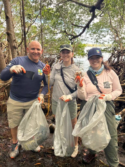 Three people are outdoors in a forested area holding trash bags and picking up litter. They are wearing gloves and casual clothing, smiling towards the camera. The background includes trees and a glimpse of water.
