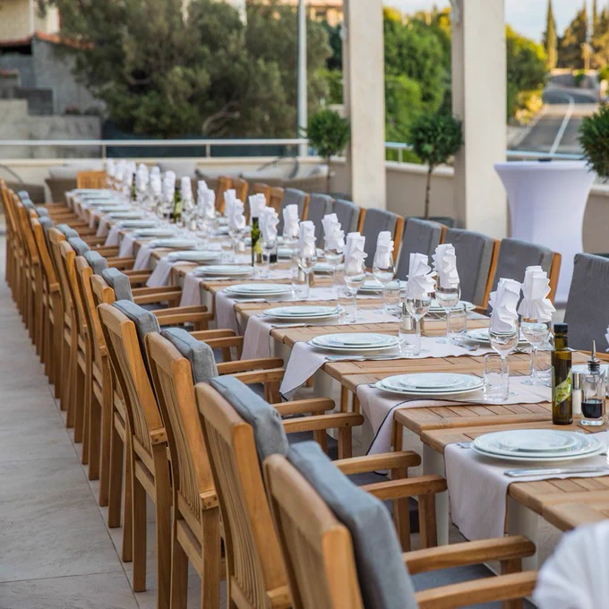 Outdoor dining area with long wooden tables set for an event, featuring white plates, glasses, folded napkins, and olive oil bottles.  Tavern Arka restaurant / La Strada Events venue - Dubrovnik, Croatia.