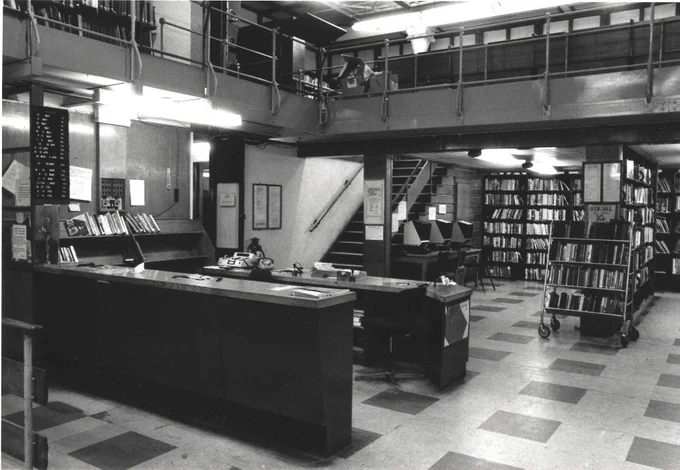 Interior of a library with circulation desk, bookshelves, and a book sale trolley.