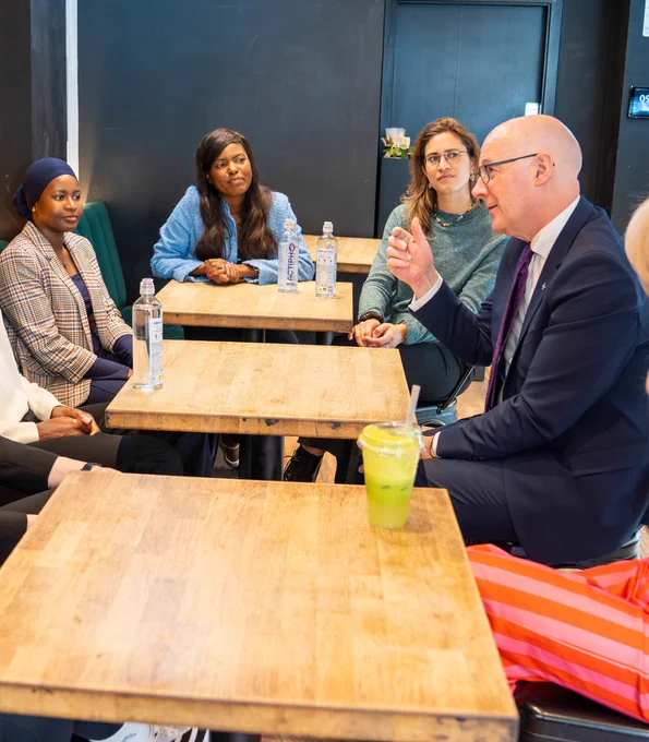 First Minister John Swinney speaking with young entrepreneurs sat around a table. 