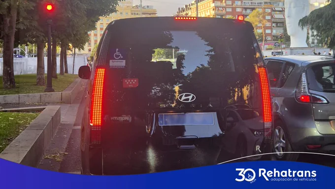 Parte trasera de furgoneta Hyundai negra con distintivo de silla de ruedas y luces traseras encendidas en semáforo.