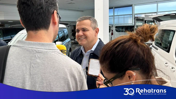 Hombre sonriente atiende a periodistas con micrófono y móvil en un concesionario de furgonetas; logo Rehatrans abajo.