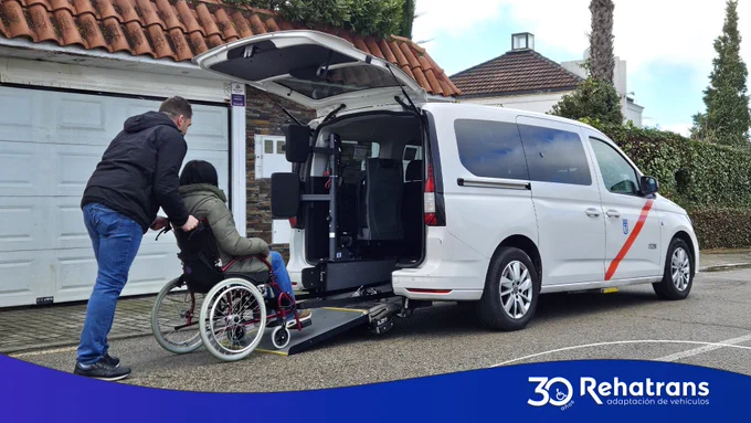 Persona en silla de ruedas sube por la rampa trasera a una furgoneta adaptada, asistida por otra persona.
