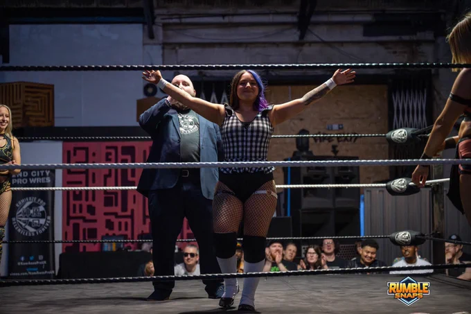An image of Kamikaze Pro ring announcer Colin Russell-Ames introducing Hollie Barlow at the beginning of a match, in the wrestling ring. Hollie is in the foreground posing, with Colin just behind her delivering the introduction into a microphone. Hollie’s opponents for the match, Aluna and Chantal Jordan, can be seen on the extreme left and right of the image respectively, in the corners of the ring.