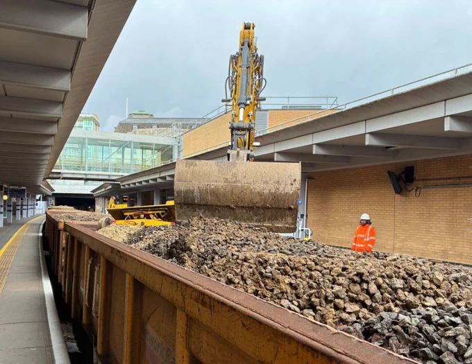 At Woolwich Arsenal station platform, a large excavator bucket is lifting and unloading ballast stones into an open freight wagon. The wagon is partially filled with grey and brown rocks. A worker in orange high-visibility clothing and a hard hat stands beside the wagon. 