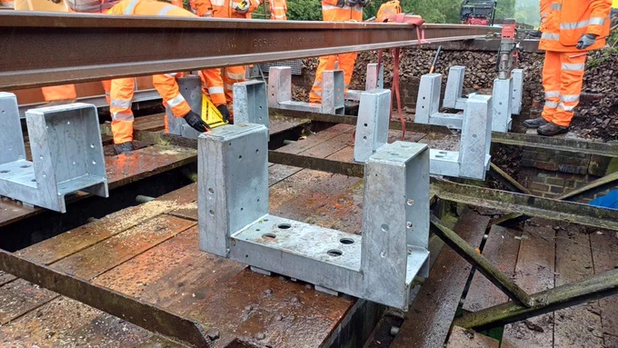 Close up of a railway bridge deck under construction in wet, rainy conditions. Multiple metal mounting brackets are fixed onto wooden beams, with standing water visible on the surface. Several workers in orange high-visibility clothing stand around the structure, preparing and aligning steel components. A long steel beam runs across the top of the frame.