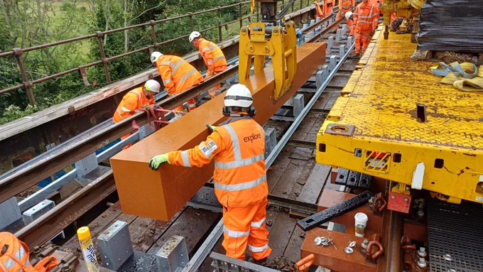 Workers in orange high-visibility clothing install a large steel beam on a bridge using a yellow lifting machine. The beam is being carefully positioned onto newly fitted supports, with several workers guiding it into place along the track. 