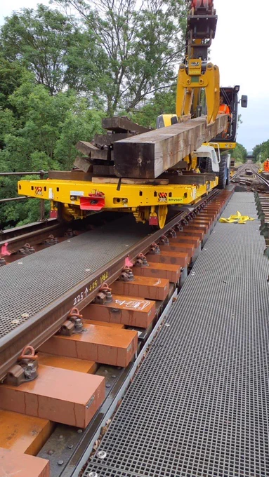 A Road Rail Vehicle (RRV) carries stacked dark wooden timbers along a track on a bridge. A crane attachment is positioned above the load, ready to lift one beam. Newly installed sleepers and rails are visible beneath, with metal safety walkways on either side. 