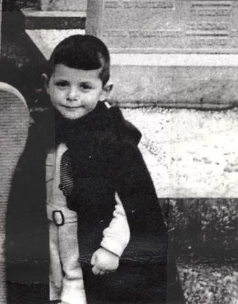 Black and white photo of a young child in a trench coat, standing confidently with one hand tucked into their pocket. The background features a partially visible engraved stone slab.