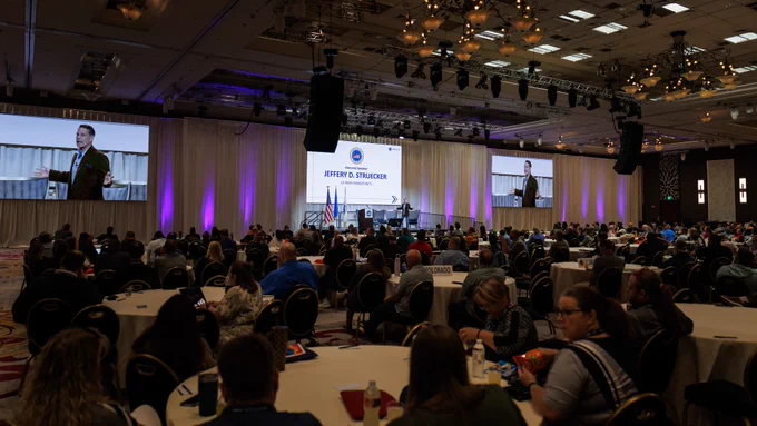 A speaker addresses a large audience at a conference. The event features live projections of the speaker and different state representatives.