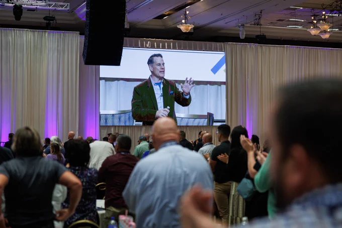 A speaker addresses an audience at a conference, with a large screen projecting his image. The atmosphere is focused and engaged.