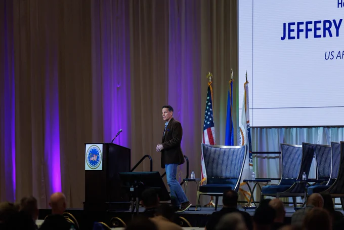 A man stands next to a podium on stage at a conference. The stage features flags and a large screen with text.