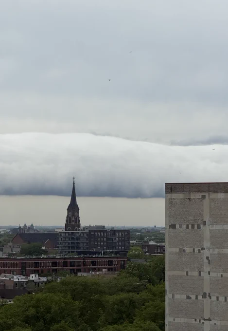 A view of a cityscape featuring a prominent church steeple, surrounded by various buildings. A large, thick cloud move across the sky, creating a dramatic backdrop. Trees and urban structures fill the foreground.