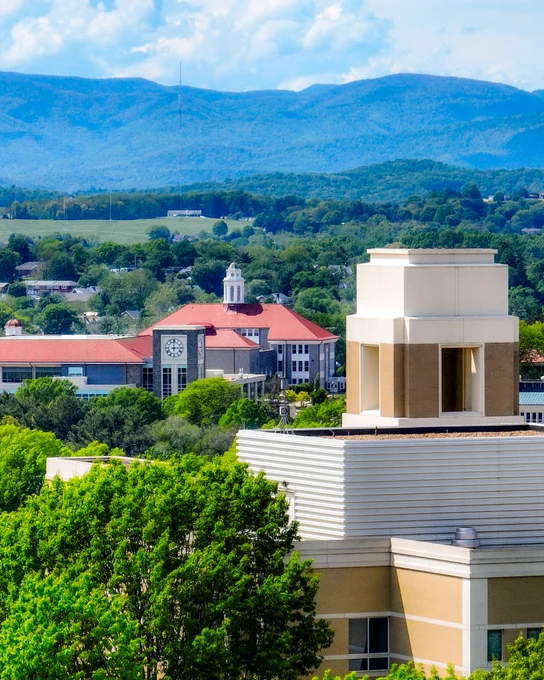 Scenic view of JMU's campus with a red-roofed bluestone buildings, including a clock tower, surrounded by trees, and a tan building in the foreground, with blue mountain ridges and scattered clouds in the background.