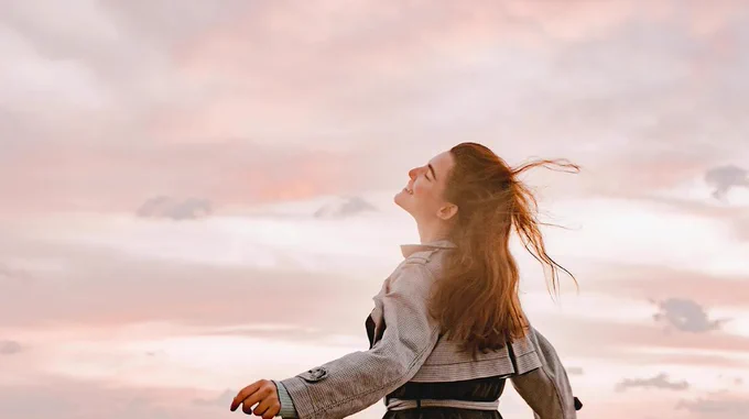 An individual with long hair stands outdoors, facing upwards with their eyes closed against a backdrop of a colorful, cloudy sky during sunset.
