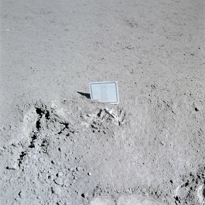 Plaque commémorative photographiée à la surface de la Lune durant Apollo 15. La plaque est partiellement enfoncée dans le régolithe et porte les noms de quatorze astronautes et cosmonautes décédés. À proximité se trouve la petite statuette Fallen Astronaut, déposée par David Scott en hommage aux pionniers disparus de l'exploration spatiale.