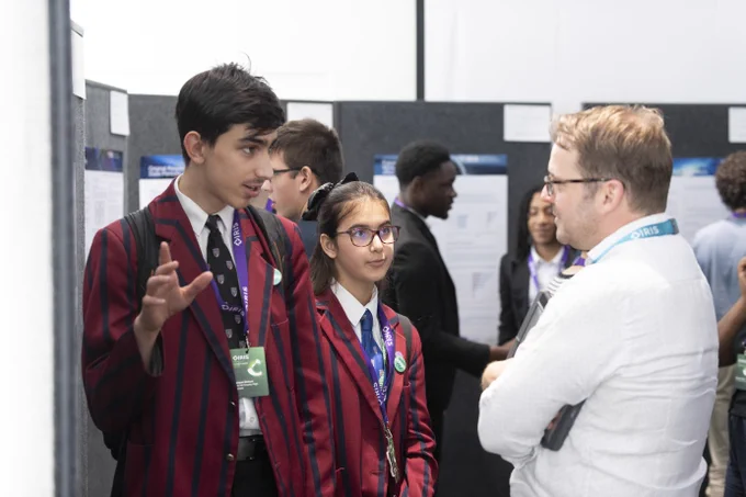 Two students stand in front of a scientific poster, speaking about their work to other people