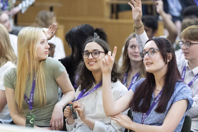 A group of students sit smiling in a conference venue. Some have their hand raised.