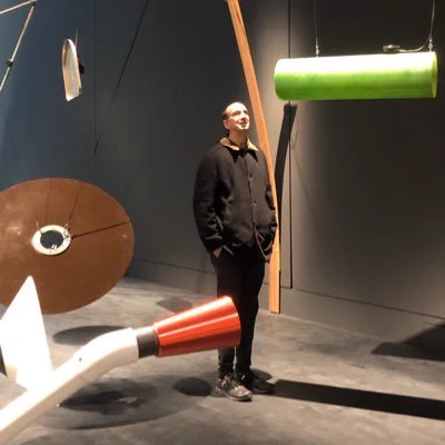 Erik Seidel, a tall, lean, balding white man, stands with his hands in his pockets. He is wearing a brown jacket and surrounded by large, colorful objects in what appears to be a museum or gallery.