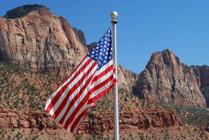 An American flag waves in the wind against a backdrop of red rock cliffs and clear blue sky in Zion National Park.