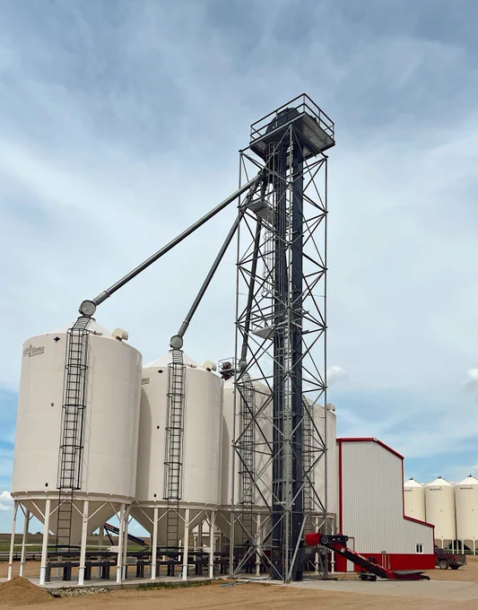 A tall grain elevator with four large white silos is connected by a metal framework on a farm. The sky is partly cloudy, and there's a small red and white structure at the base.