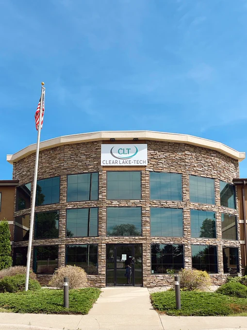 A round, three-story building with a stone facade and large glass windows under a clear sky. A sign on the building reads "Clear Lake Tech." An American flag is on a pole in front of the building, surrounded by trimmed bushes and a concrete pathway.