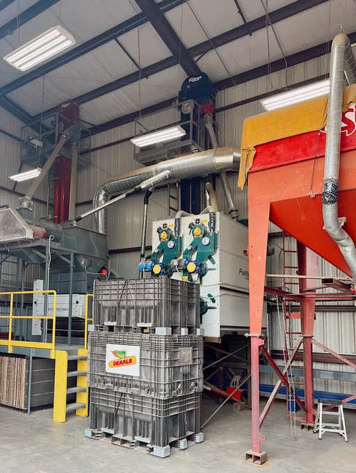 The image shows industrial equipment inside a facility, including a Dekalb-branded container in the foreground. There are large red and gray machines with metal pipes and a tall ceiling with overhead lights.