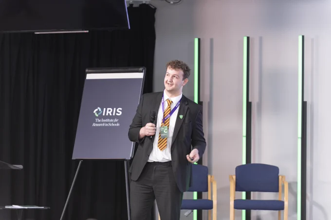 A student stands on stage holding a remote, speaking to an audience at a conference